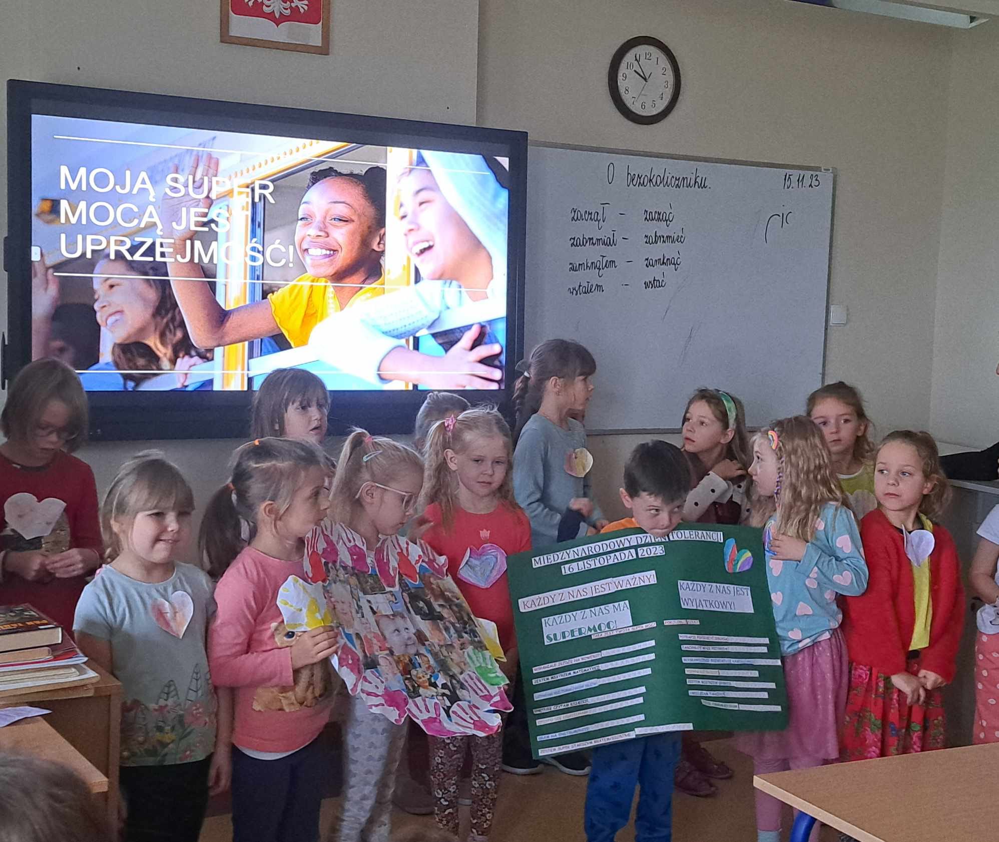 Dzieci z plakatem stoją na tle włączonej tablicy interaktywnej.
