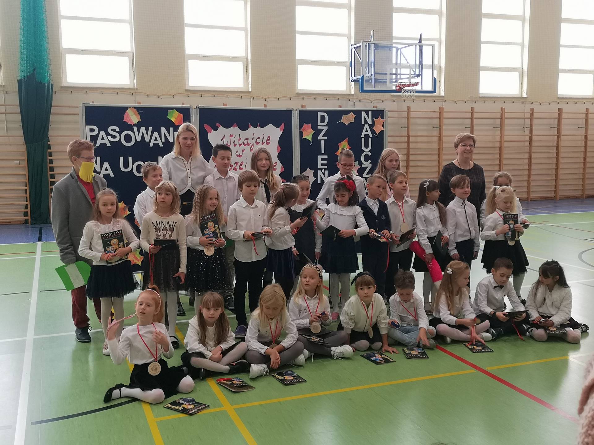 Uczniowie klasy pierwszej podczas pasowania na ucznia stoją w grupie, za nimi stoją uczniowie z klasy czwartej, nauczycielki. 